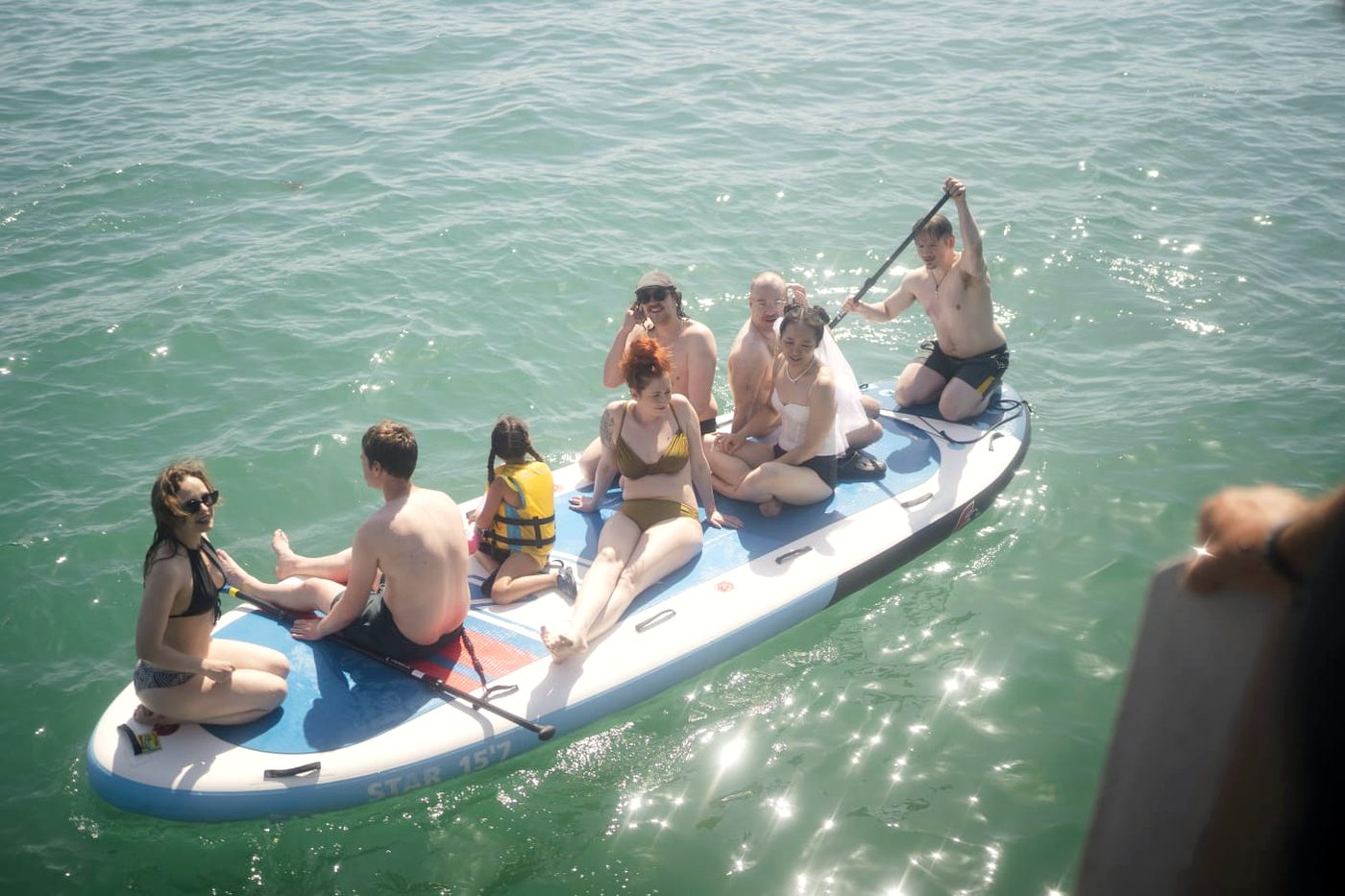 Sechs junge Leute sitzen gemeinsam auf einem Big SUP im sonnigen Wasser