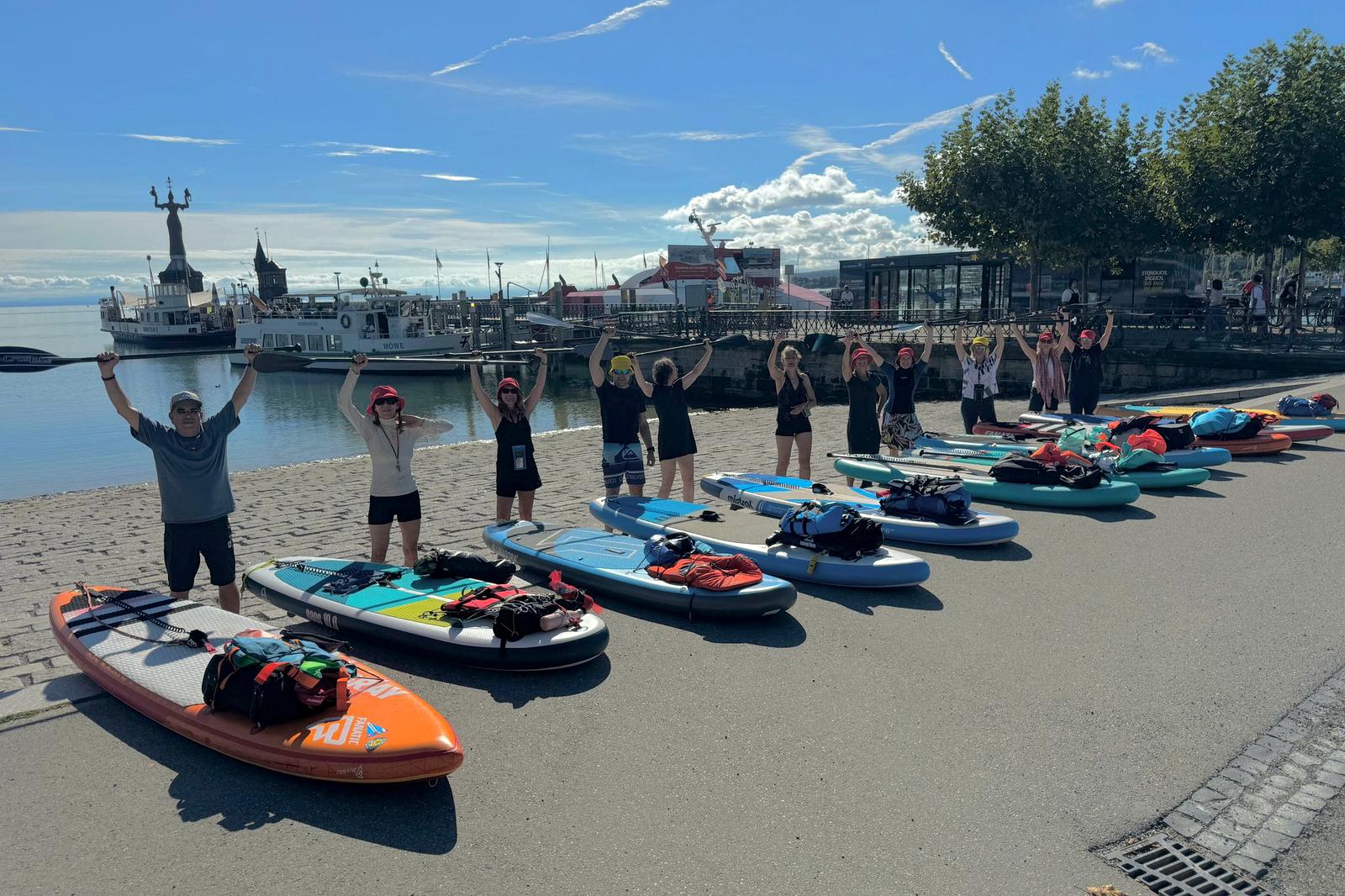 Kursgruppe steht mit erhobenen Armen am Strand hinter einer Reihe bunter SUP-Boards